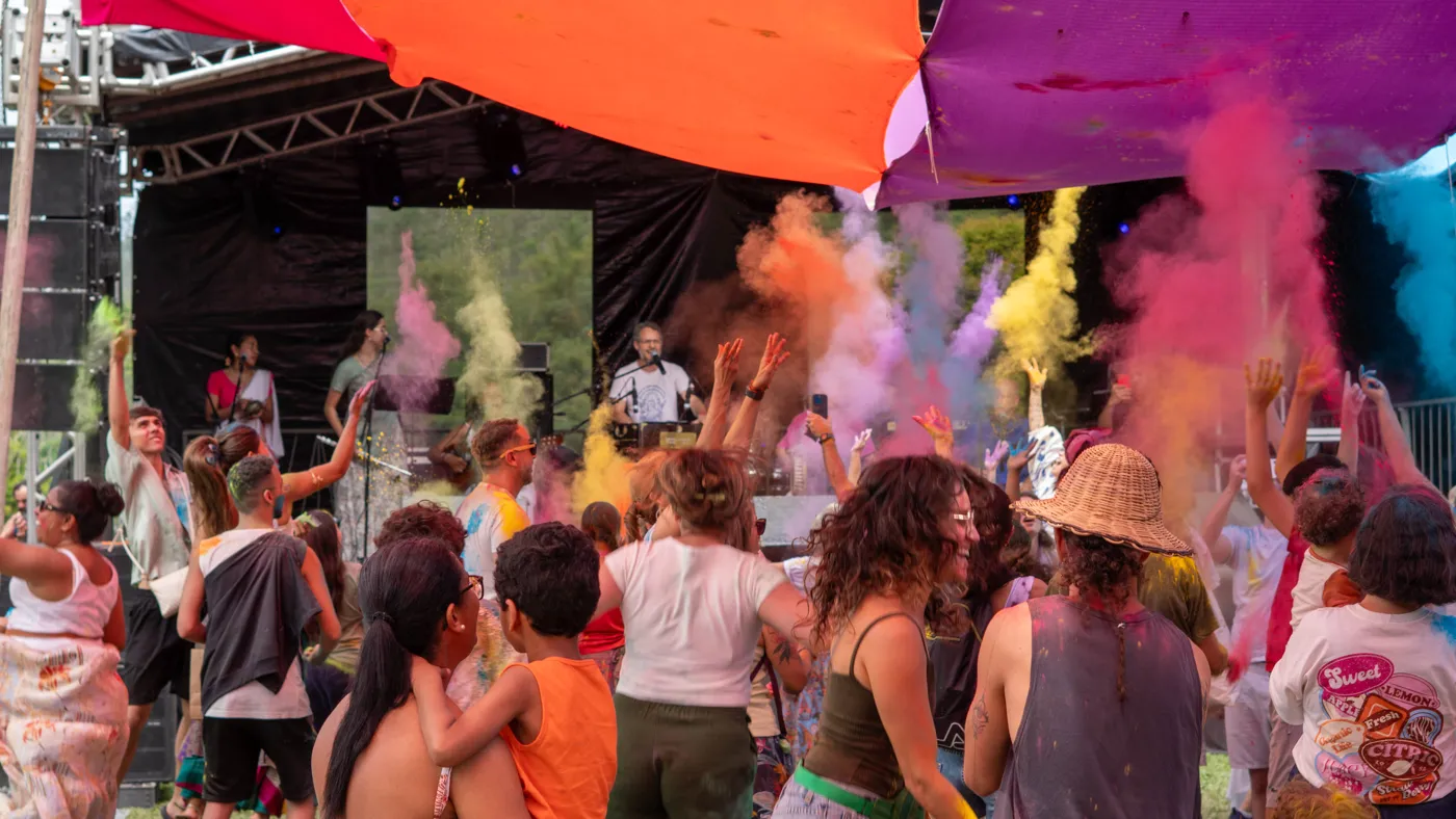 Público em festa sob tecidos laranja e lilás com pó colorido no ar