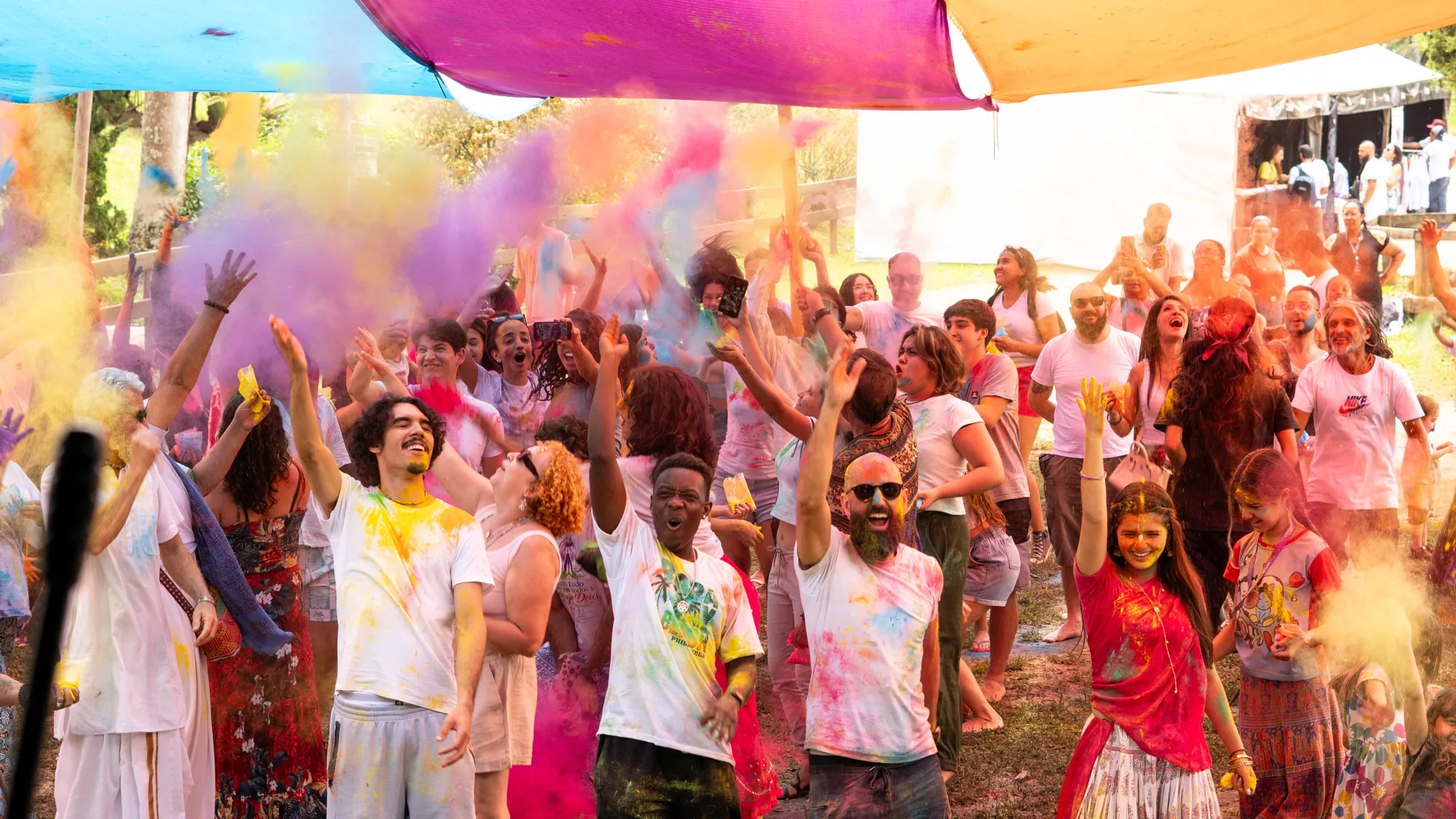 Público diverso de várias idades erguendo os braços sob tecidos coloridos enquanto nuvens de pó amarelo, rosa e lilás explodem no Holi 2026 na Fazenda Nova Gokula